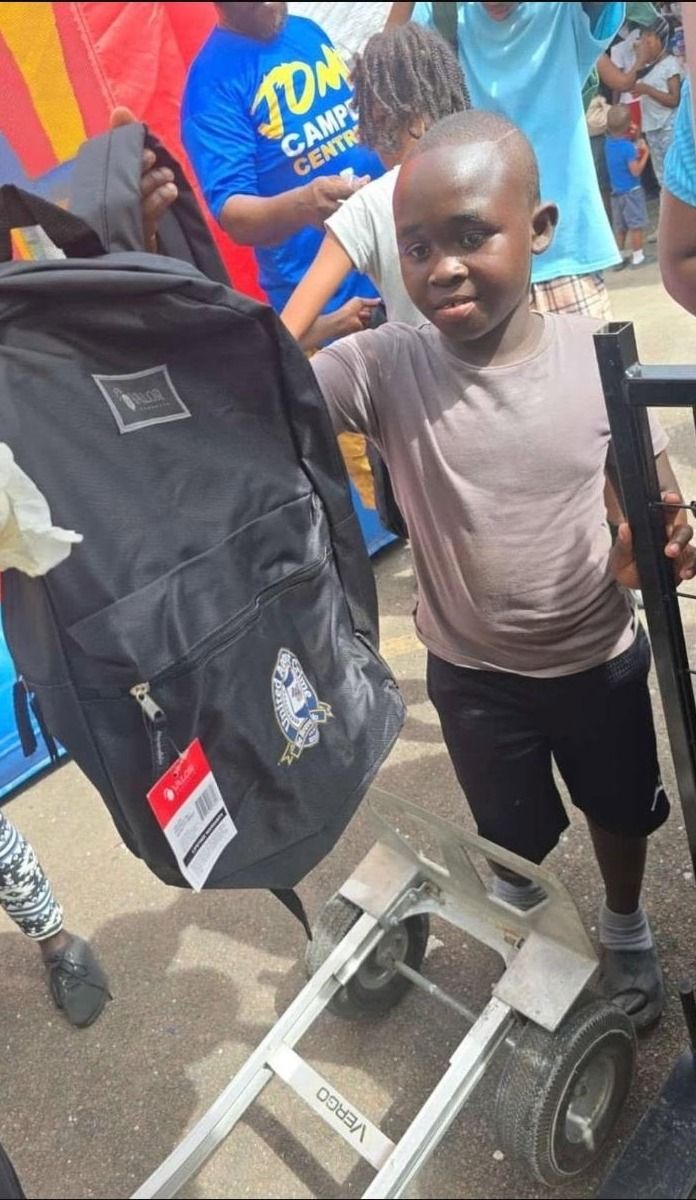 A boy stands beside a hand truck holding a new black backpack with a school crest