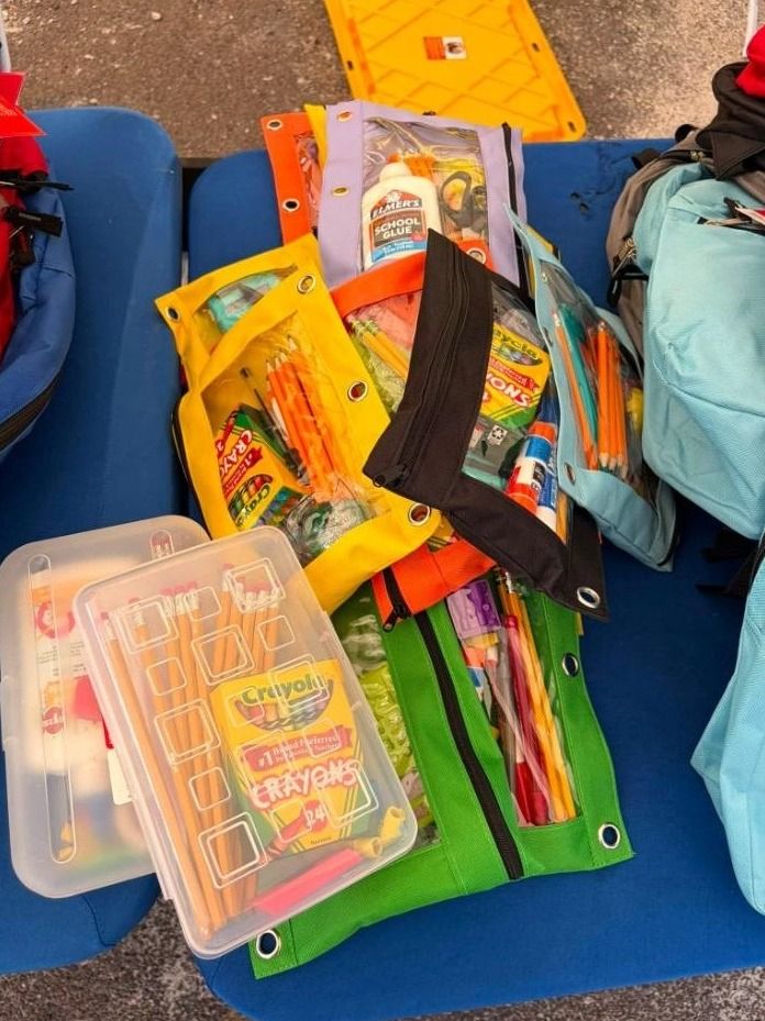 School supply pouches holding glue, crayons and pencils laid out on a blue table beside backpacks