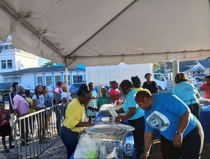 Volunteers in light blue shirts serve plates of food to a line of people under a tent