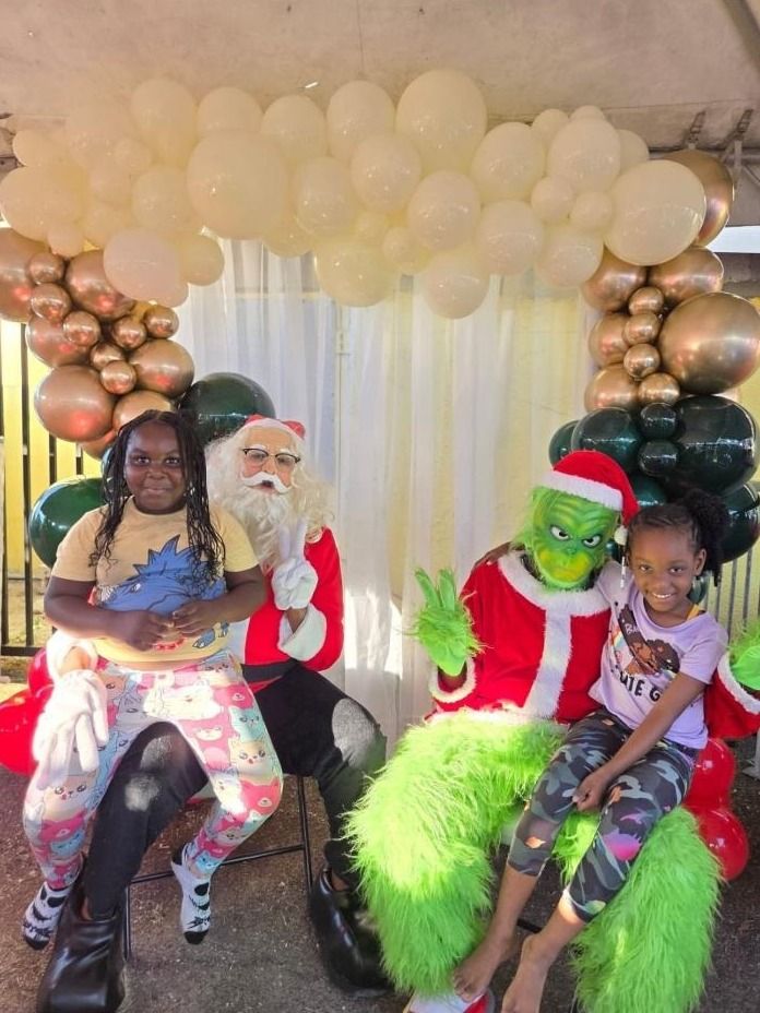 Two girls pose with Santa and a costumed Grinch in front of a cream, gold and green balloon arch
