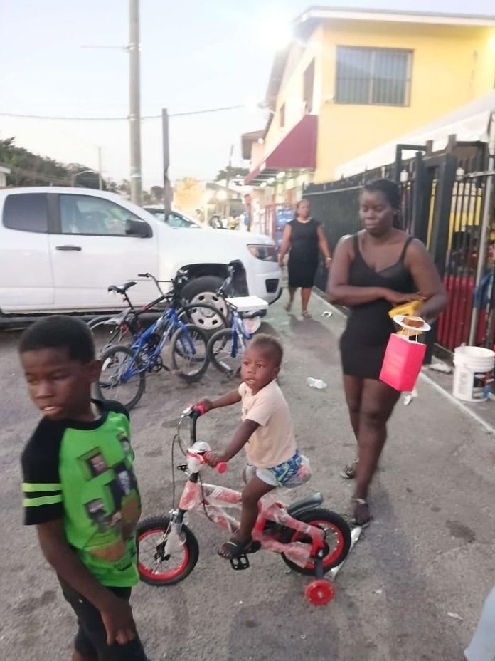 A toddler rides a small bicycle with training wheels as a woman with a pink gift bag walks past