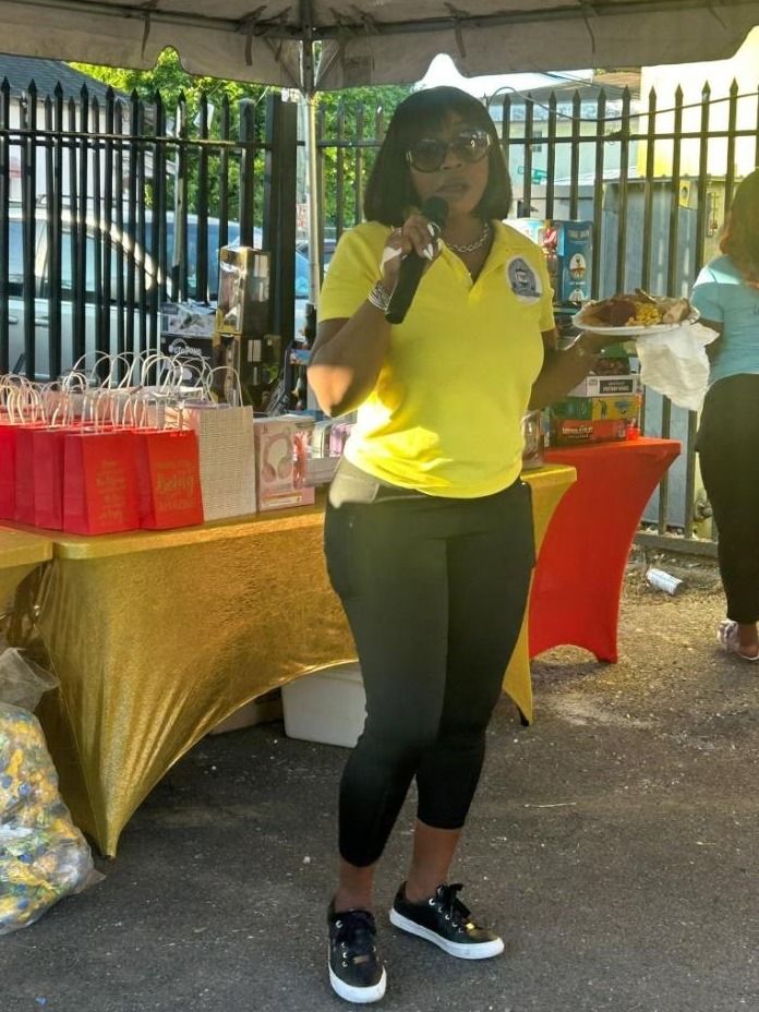 A woman in a yellow polo speaks into a microphone beside tables of red gift bags and toys