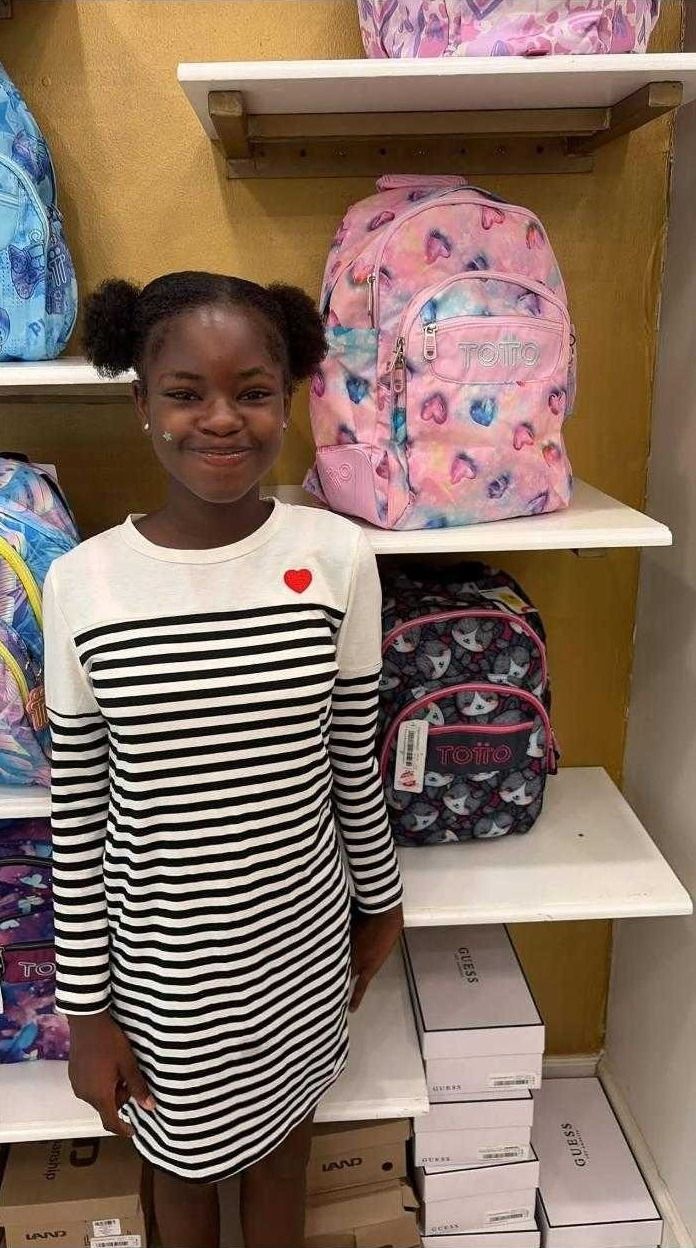 A smiling girl in a striped dress stands beside shelves of school backpacks in a store