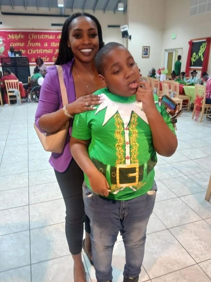 A woman in a purple blazer poses with a boy in an elf shirt in a hall decorated for Christmas