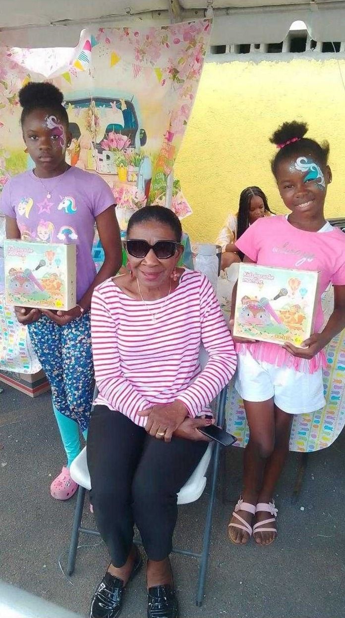 Two girls with face paint hold boxed kids karaoke machines beside a seated woman in sunglasses