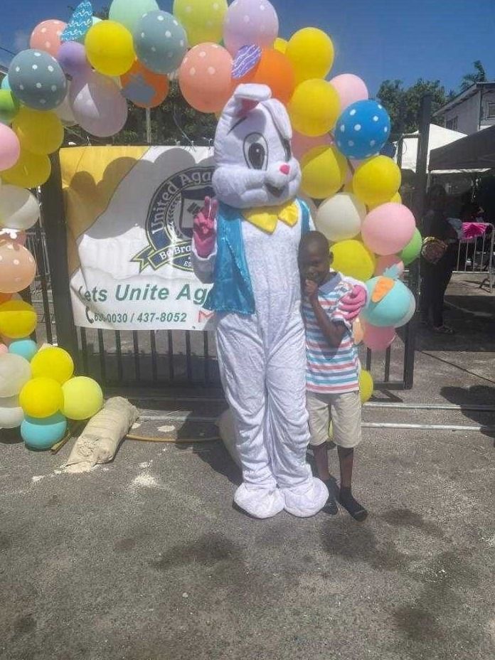 A boy stands beside a costumed Easter Bunny in front of a United Against Crime banner and balloons