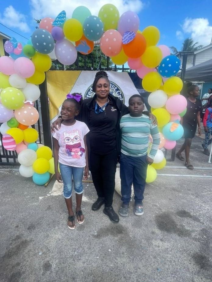 A woman in black poses with a girl and a boy in front of a pastel balloon arch