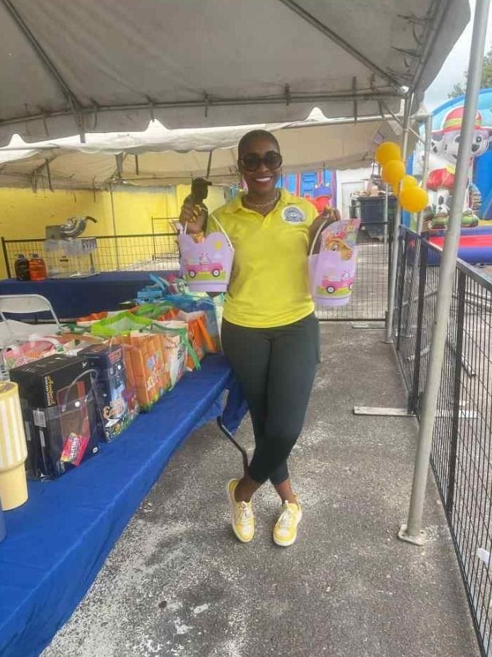A woman in a yellow polo holds two Easter gift buckets beside tables of prizes under a tent