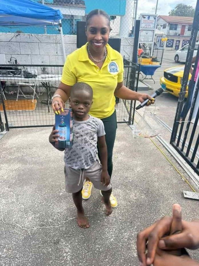 A woman in a yellow UAC polo holding a microphone stands with a barefoot boy holding a drink