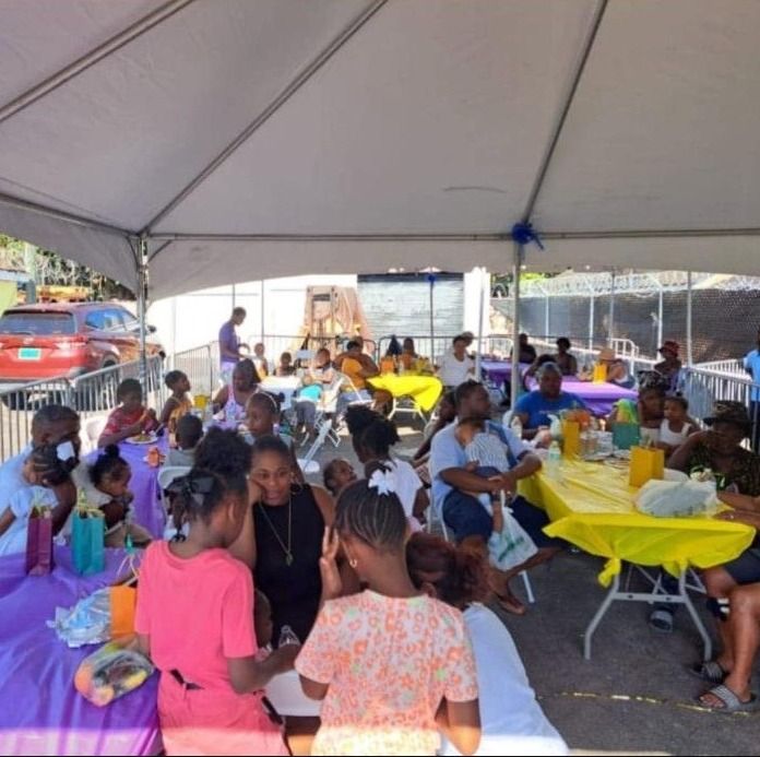 Children and adults sit at tables with yellow and purple cloths under a large tent