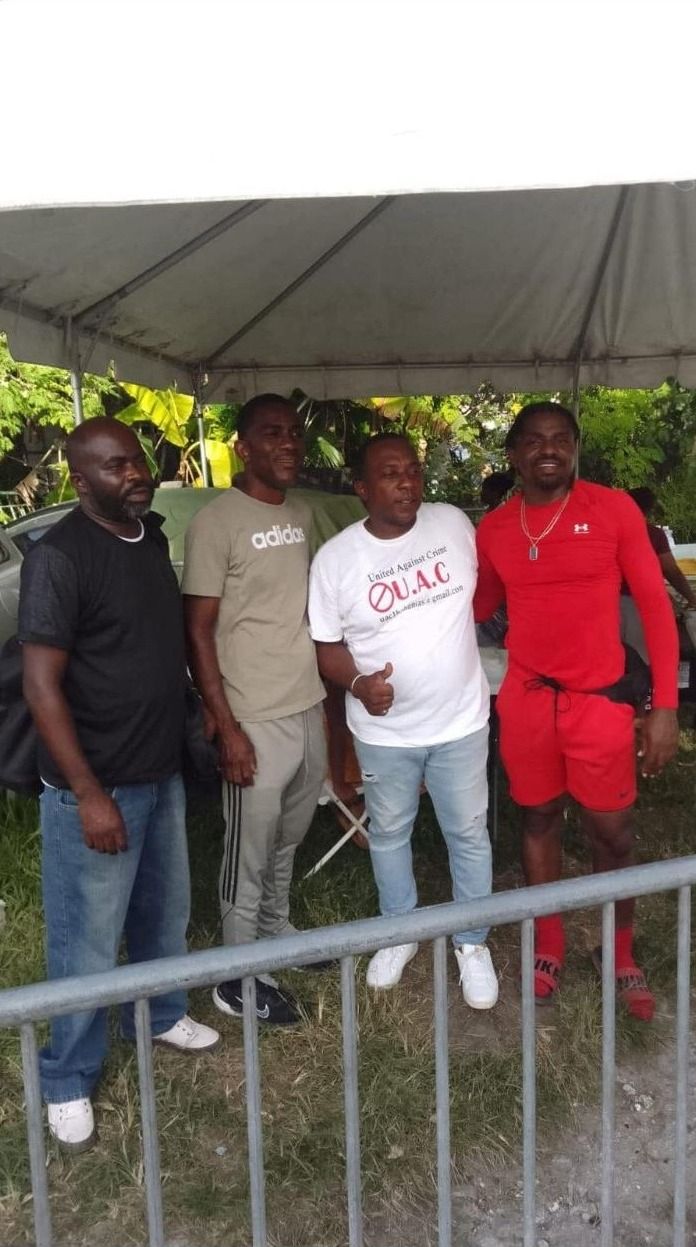 Four men pose together under a tent, one in a white United Against Crime shirt giving a thumbs up