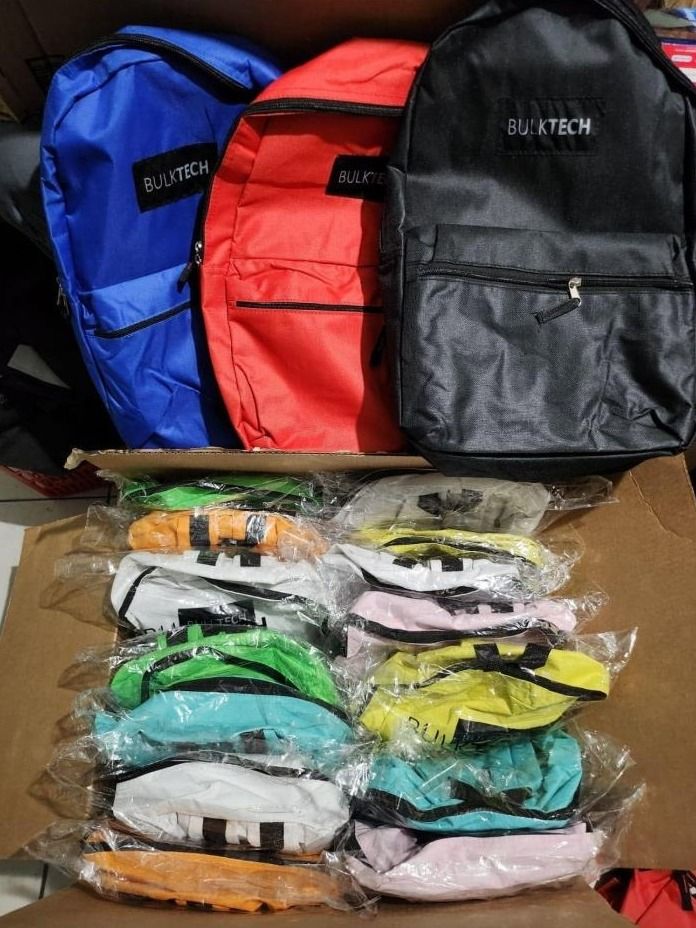 Blue, red and black backpacks sit above a box of smaller bags in clear wrap