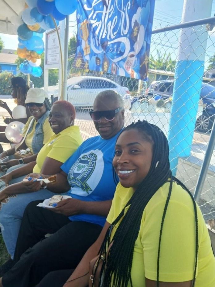 Smiling volunteers in yellow and blue shirts sit in a row eating cake beside a Father's Day balloon arch