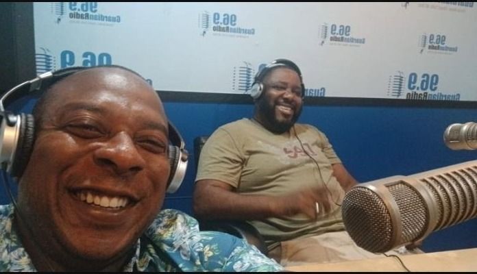 Two men in headphones smile beside a large microphone in the Guardian Radio 96.9 studio