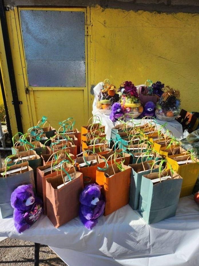 Paper gift bags, purple plush bears and fruit baskets are arranged on a white table against a yellow wall