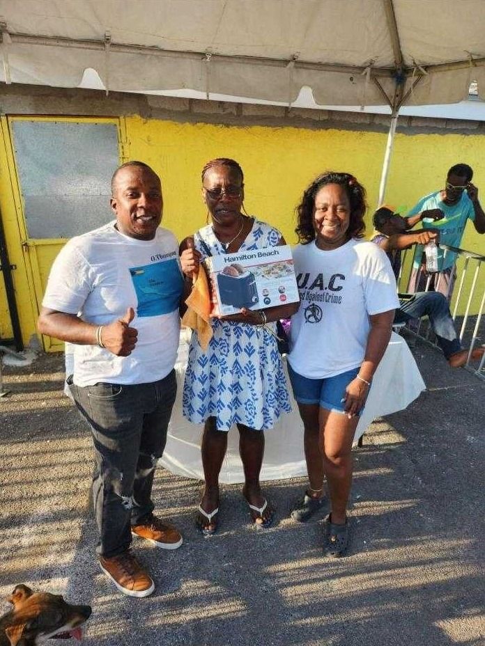 Two volunteers in white shirts present a woman in a blue patterned dress with a boxed toaster
