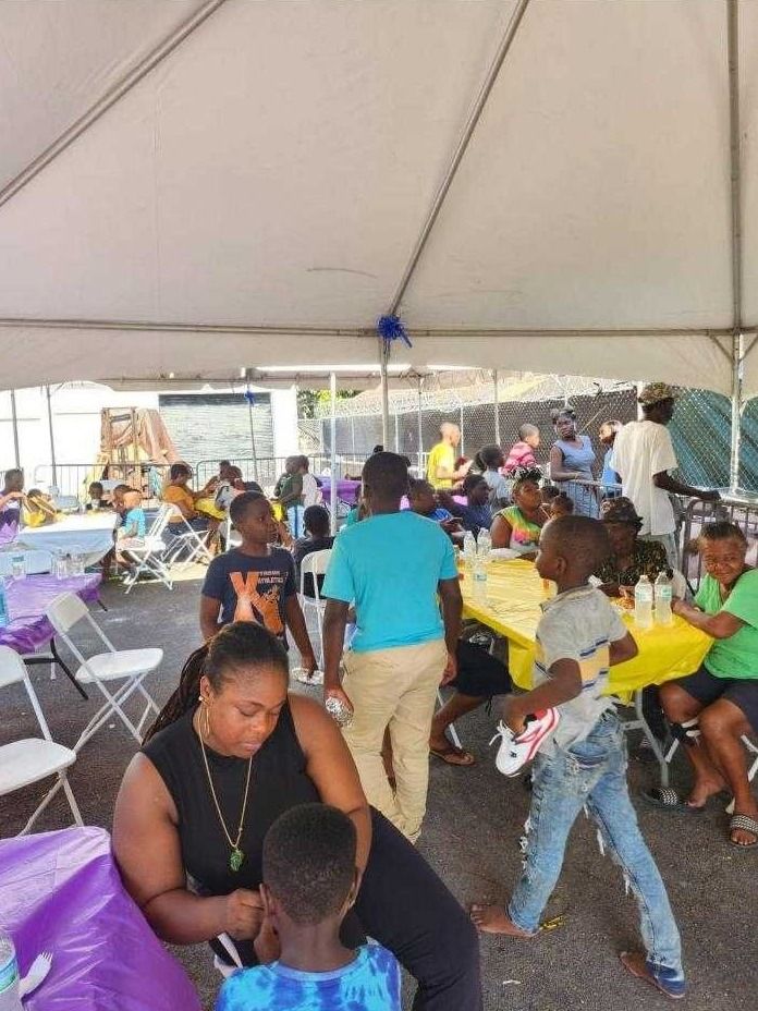 Children and adults sit at tables with yellow and purple cloths under a large tent