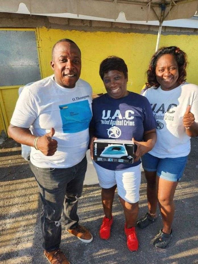 Two volunteers in white shirts give a woman in a navy U.A.C. shirt a boxed iron, all smiling