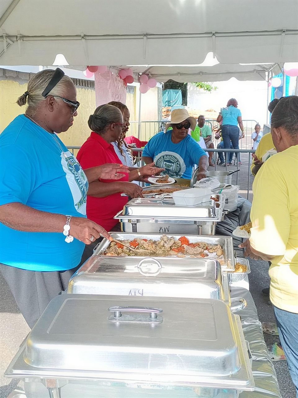 Volunteers in blue and red shirts serve hot food from chafing dishes under a tent