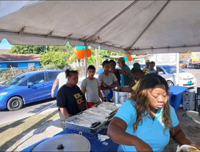Volunteers serve food from foil trays and a large pot to people waiting in line under a tent