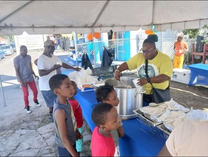 A woman in a yellow shirt stirs a large pot of food while children wait at the blue serving table